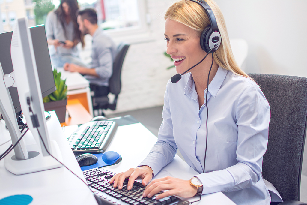 Hymyilevä naispuolinen asiakaspalvelija kuulokkeilla tietokoneen ääressä
